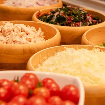 木の器に盛られた、細切りにした鶏肉、シュレッドチーズ、ミニトマトなどのさまざまなサラダバーの具材のクローズアップ写真。