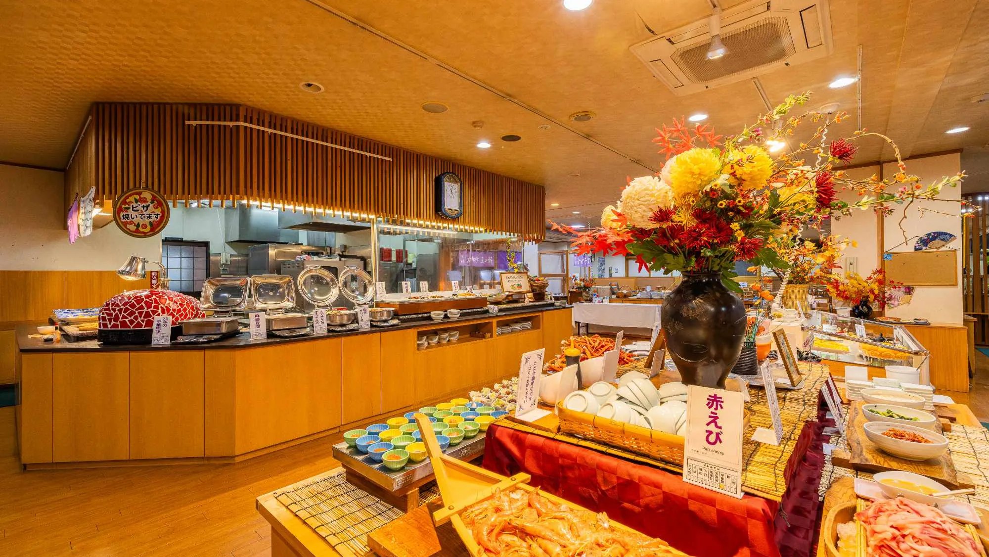 ビュッフェ形式の料理が並ぶ、明るく広々としたバイキング会場。手前には大きな花瓶に生けられた豪華な生花があり、その周囲に様々な料理や器が並んでいる。