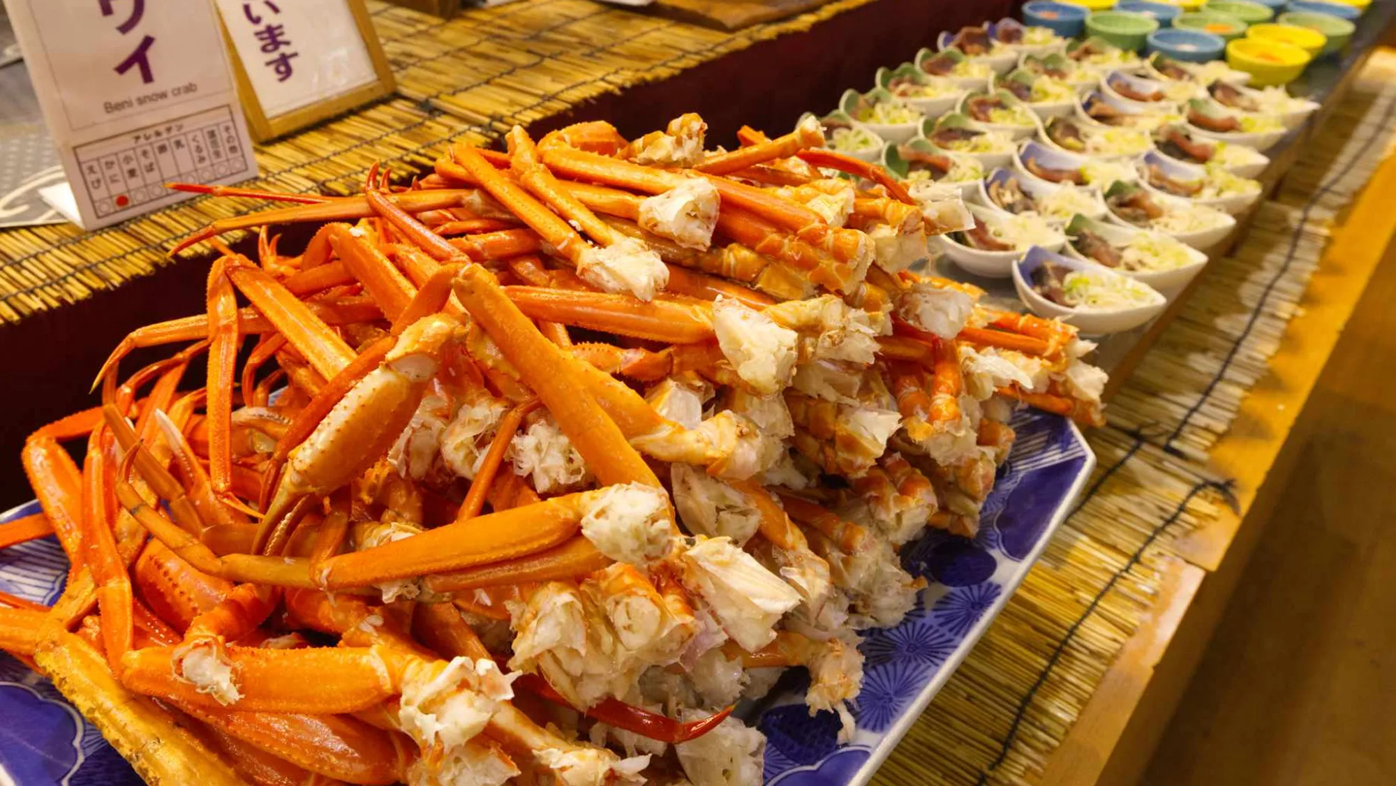 ビュッフェのテーブルに山積みにされた、茹でられたベニズワイガニの脚。鮮やかなオレンジ色で、一部は殻が割られて白い身が見えている。その奥には、一口サイズのオードブルのような小皿料理が整然と並べられている。