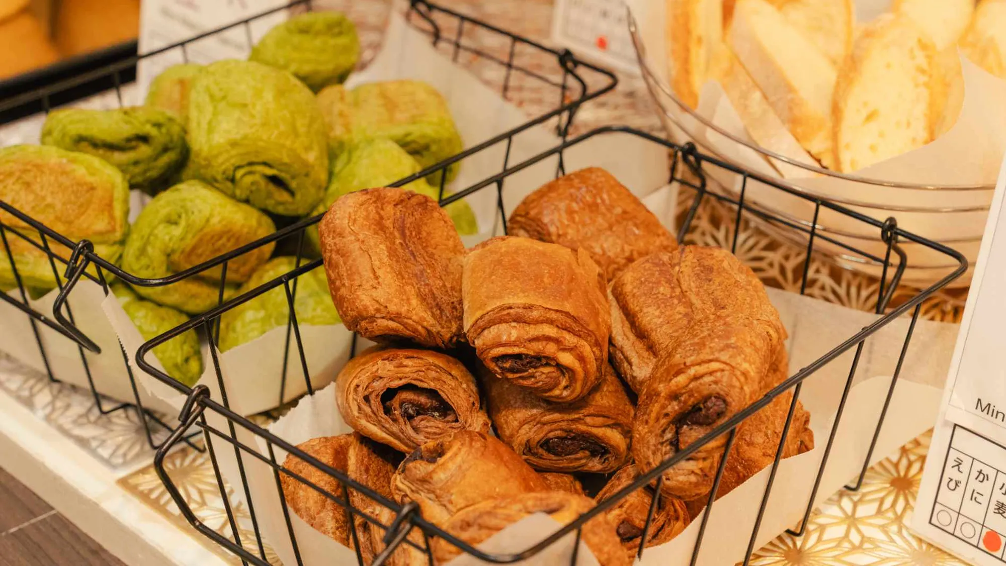 黒いワイヤーバスケットに盛られた焼き立てのパン。手前には茶色く焼き上がったチョコレート入りのデニッシュが、左奥には鮮やかな緑色をした抹茶風味のデニッシュが並んでいる。