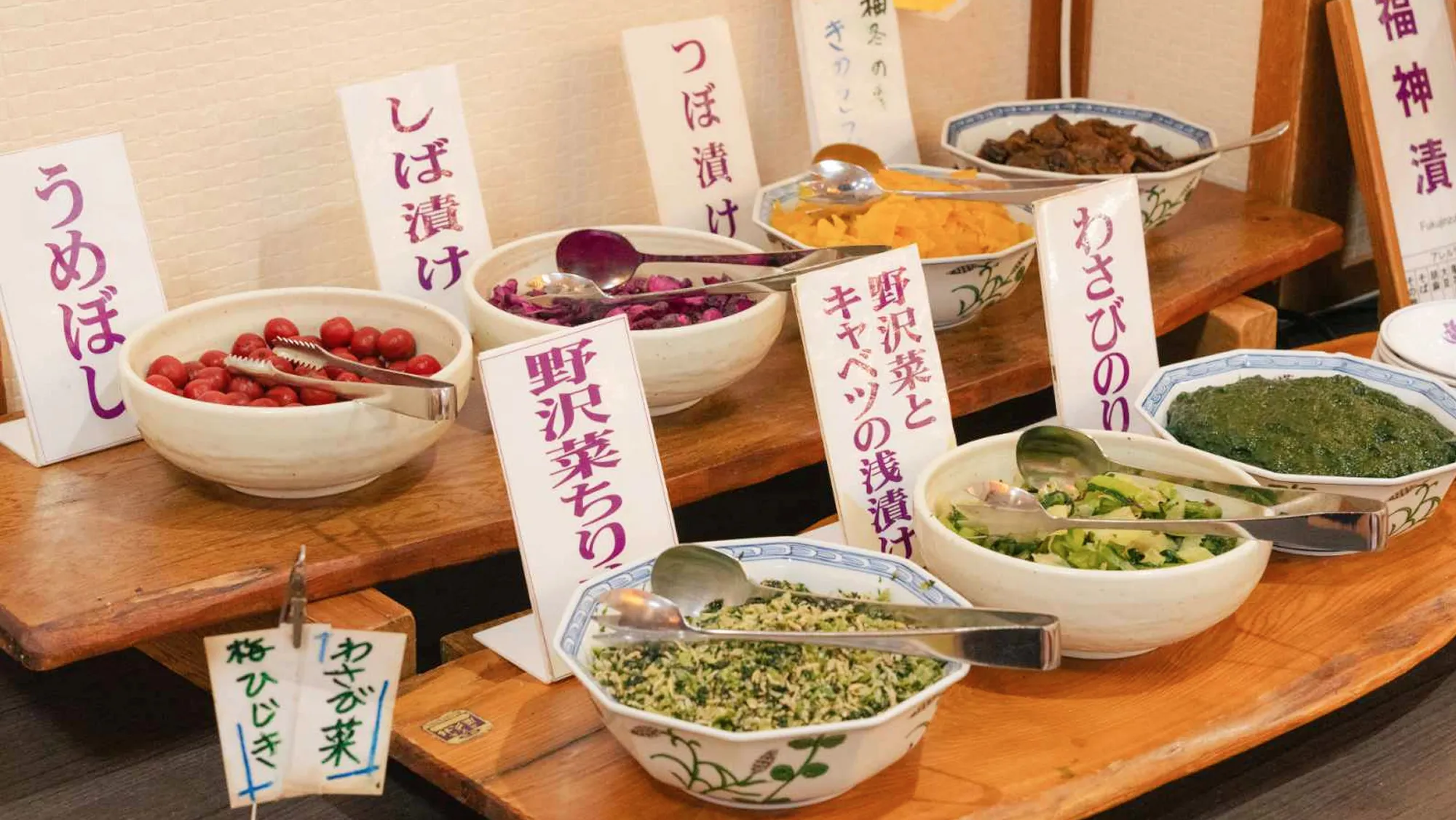 二段になった木製の台の上に、様々な種類の和風の漬物やご飯のお供が白い陶器の鉢に盛られている。手前の鉢には「わさび菜」や「野沢菜ちり」、奥には「うめぼし」「しば漬け」「つぼ漬け」などの札が添えられている。