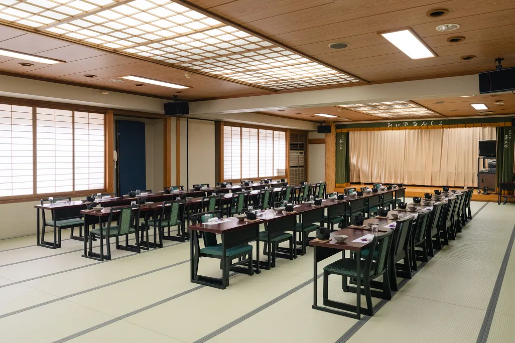 畳の床に黒い脚のテーブルと深緑色の椅子が整然と二列に並べられた広々とした大宴会場で、周囲の壁には障子のような大きな窓があり、奥にはステージと舞台幕が設置されており、大人数の宴会や式典に対応できる和風の空間である。