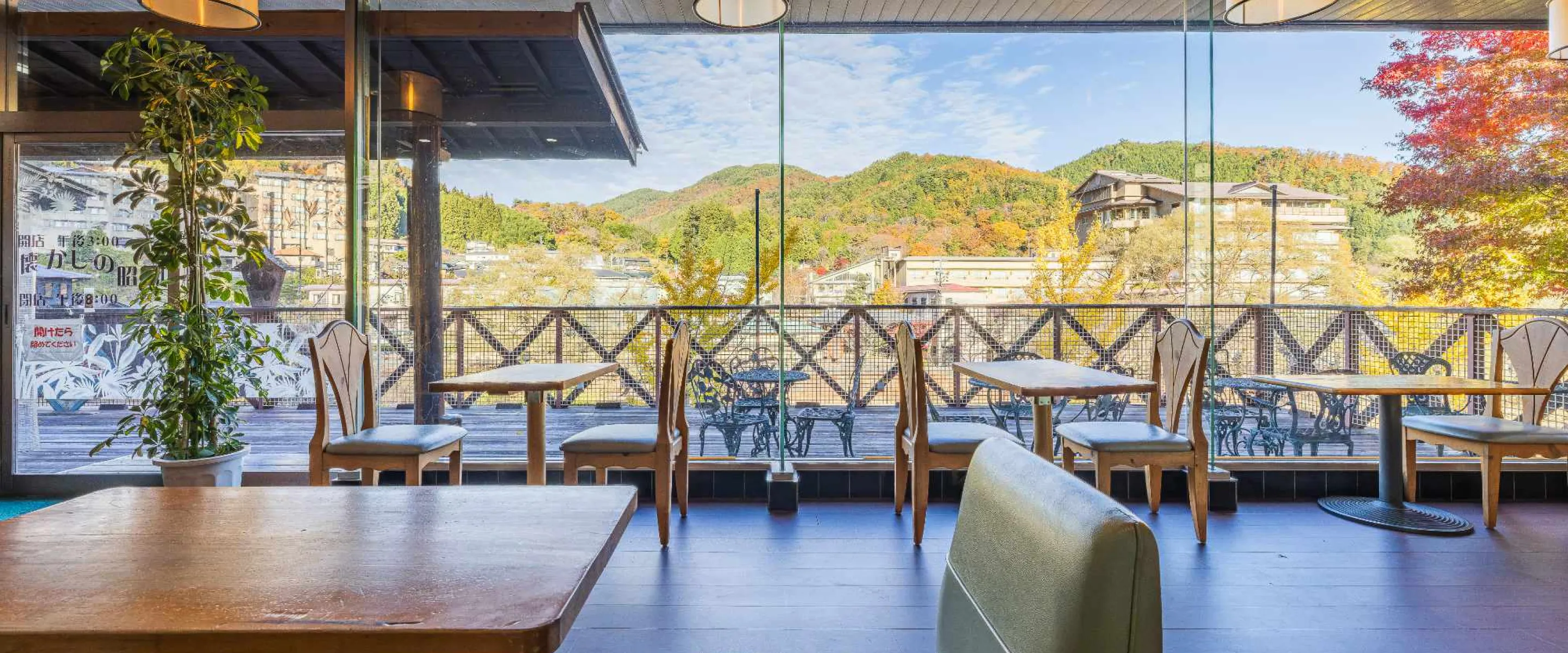 大きな窓ガラスの向こうに紅葉に色づいた山々や温泉街の景色が広がる、開放的で眺めの良い食事・休憩スペースで、特徴的な背もたれの椅子と木製のテーブルが窓際に並び、明るい日差しが差し込むリゾート感あふれる空間である。