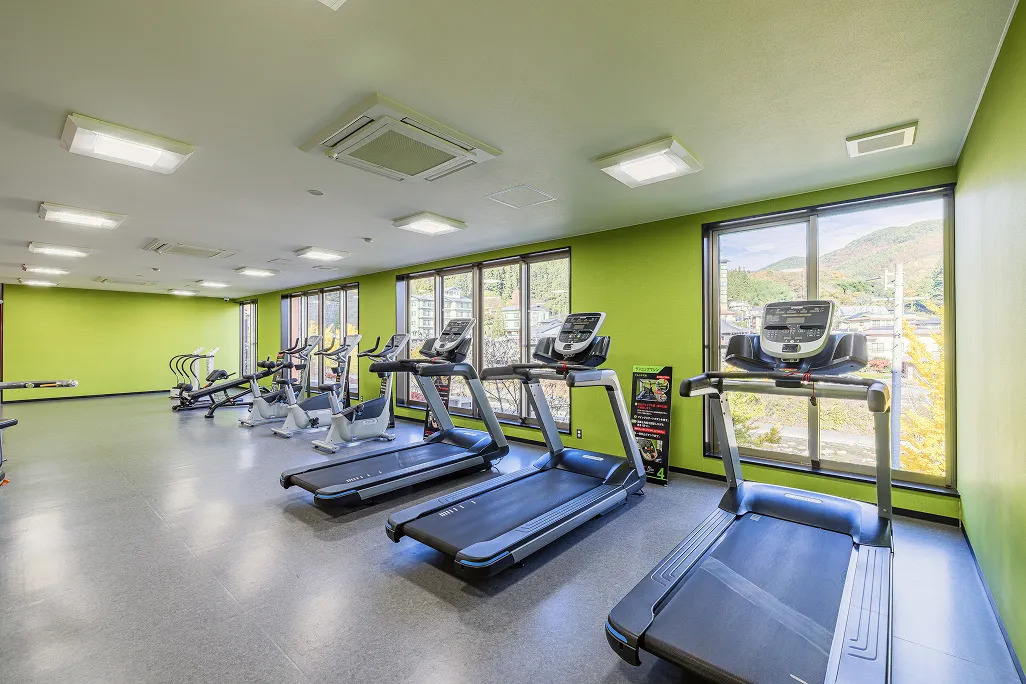 ライムグリーンの壁と大きな窓が特徴的な明るいフィットネスジムで、窓の外に紅葉した山々の景色が見える手前にはランニングマシンが3台並び、その奥にはエアロバイクやストレッチマシンなどの各種トレーニングマシンが整然と配置されている。
