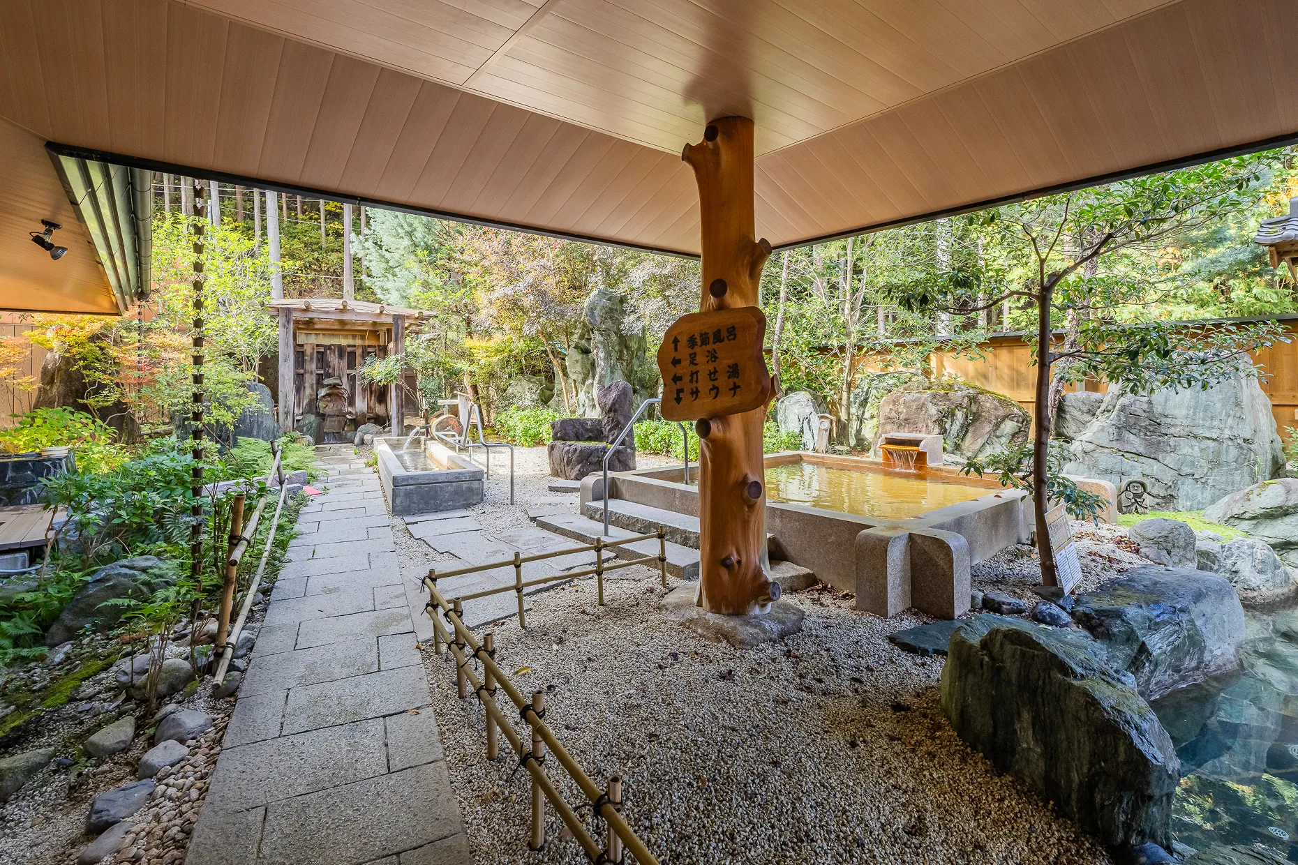 日差しが差し込む開放的な屋根の下に、木の幹の柱と岩に囲まれた二つの湯船が並び、小石と飛び石の通路や豊かな緑の木々が配された、趣のある庭園露天風呂の風景である。