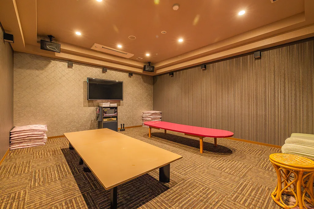 ブラウンとベージュの幾何学模様のカーペットが敷かれ、壁には音響設備と大きなテレビモニターが設置された広いカラオケルームで、中央にはローテーブル、奥にはピンクのベンチと座布団が積まれ、大人数での利用に対応した明るい空間である。