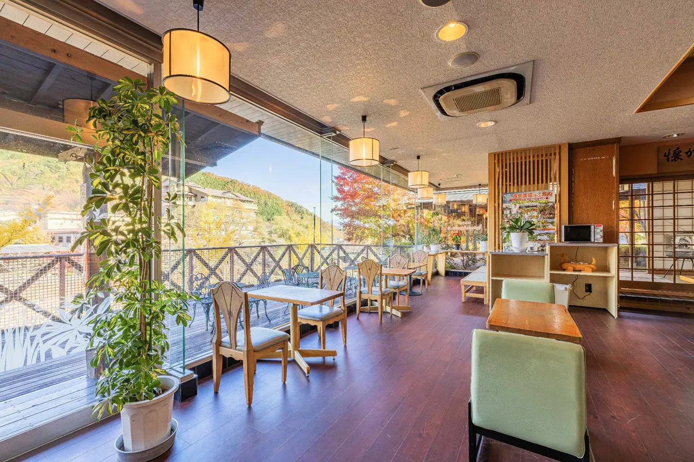 大きな窓ガラス越しに色づいた山々や温泉街の景色を一望できる開放的なロビーラウンジで、木製のテーブルと背もたれが特徴的なデザインの椅子、一部に緑のソファ席などが配置されており、明るい日差しが差し込む中でゆったりとくつろげる空間である。