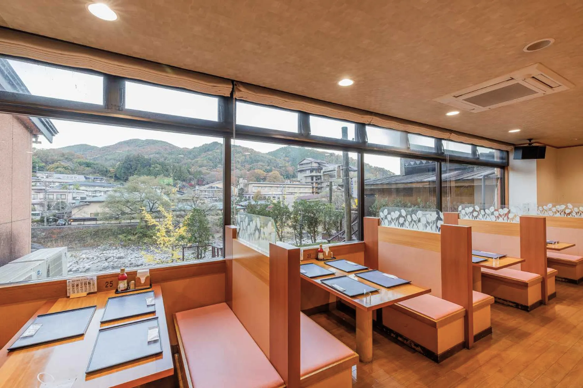 旅館の食事処の窓際の席。窓の外には紅葉の始まった山々や、川沿いの温泉街の景色が広がる。窓に沿って、オレンジがかったベージュの背もたれの高いベンチシートが並び、木製の小さなテーブルが配置されている。