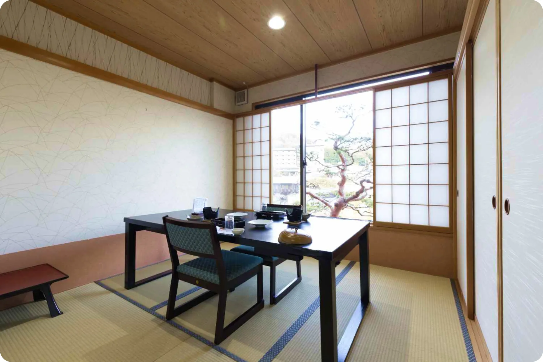 畳敷きの和室にある食事スペース。中央にはモダンな黒いダイニングテーブルが置かれ、椅子が2脚と、反対側には座布団が見える。窓際には障子があり、外には手入れされた庭の木が見える。