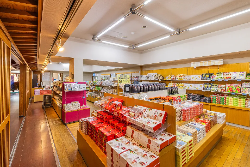 明るい照明の広い店内に、信州の銘菓や土産物が棚や台に多数陳列されたお土産処「さんぽ道」の様子で、奥の壁は明るい黄色の棚、手前は木製の床と朱色のディスプレイ台が特徴的な、活気のある空間である。