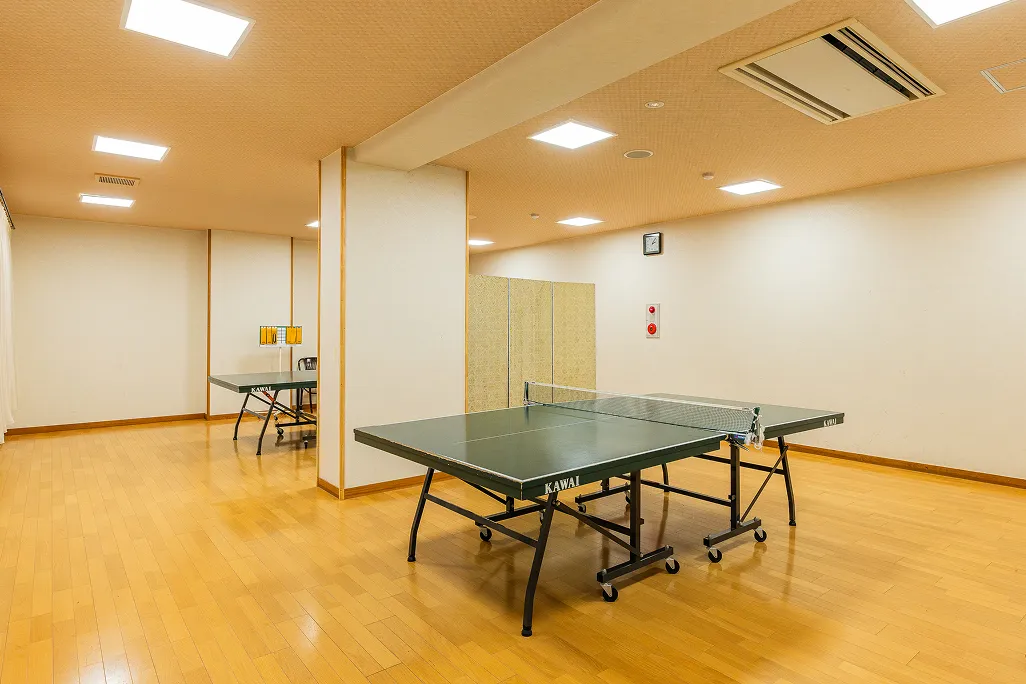 明るいフローリングの床と白い壁の広々とした卓球場で、手前に緑色の卓球台が一つ置かれ、その奥にもう一つ卓球台が見えており、湯上がりなどに気軽にアクティビティを楽しめる空間である。