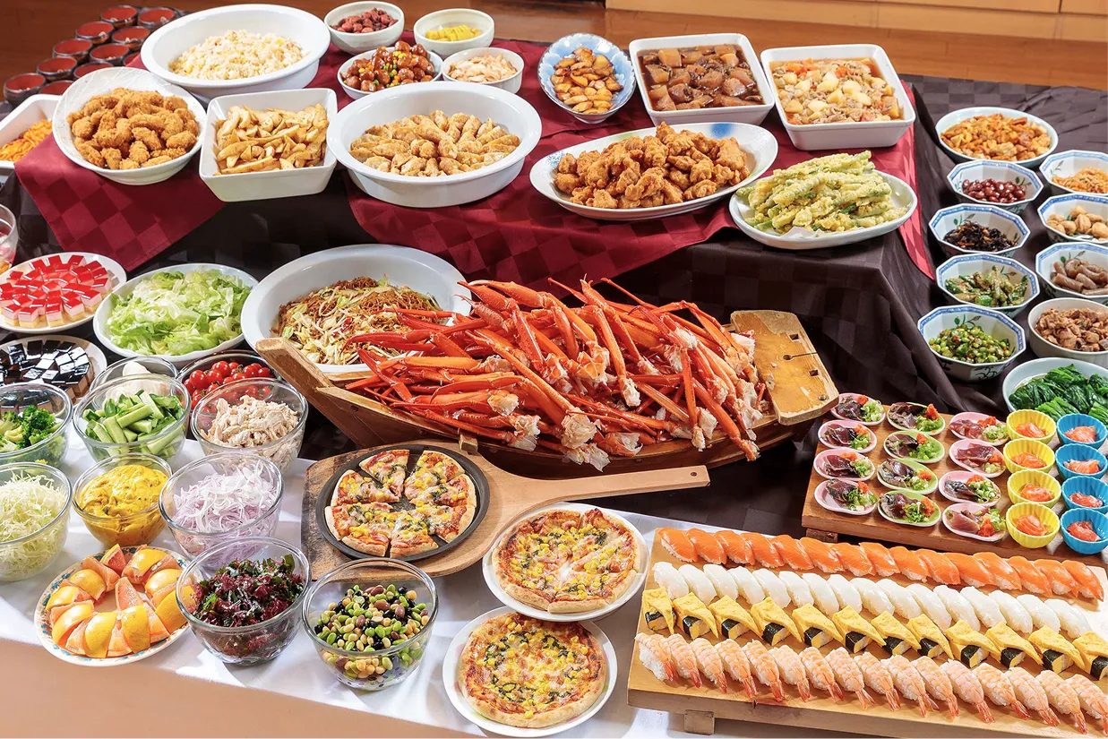 テーブルいっぱいに並んだ和洋中のバイキング。手前には握り寿司やピザ、中央には山盛りのカニ、奥には数多くの大皿料理やサラダ、デザートが所狭しと並ぶ様子。