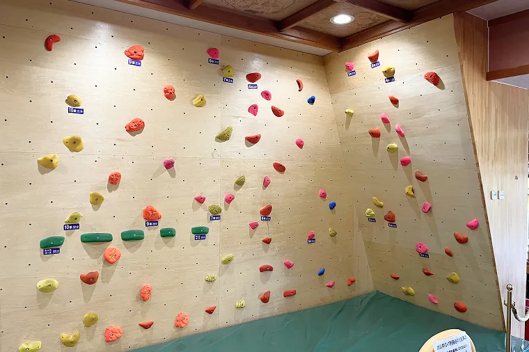 室内に設置された、カラフルなホールドが散りばめられた木製パネルのボルダリング壁。足元には安全のための大きな緑色のマットが敷かれている。