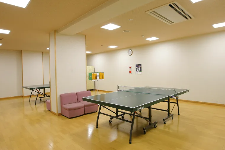 明るい照明の広々とした多目的ホールに設置された、2台の本格的な卓球台。壁際には休憩用のピンク色のソファがあり、家族やグループでゆったり楽しめる空間