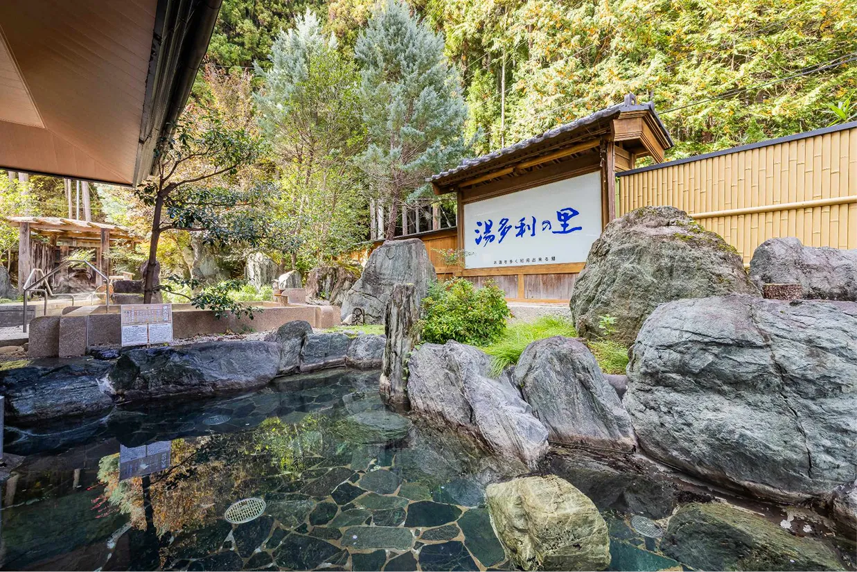 大きな岩に囲まれた開放的な庭園露天風呂。澄んだお湯の向こうには豊かな木々と「湯多利の里」と書かれた看板が見える落ち着いた空間