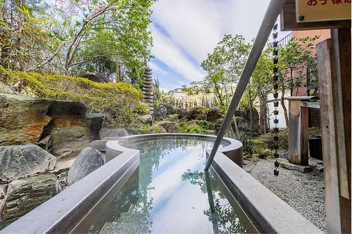 空を映し出す細長い鍵穴のような形をした、石造りの露天風呂。周囲には庭園の木々や岩が配され、落ち着いた和の風情を感じる空間。