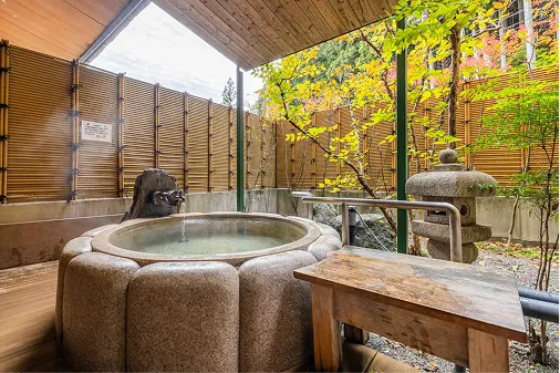 竹垣に囲まれた、一輪の花のような形をした石造りの円形露天風呂。傍らには石灯籠が置かれ、黄色く色づいた紅葉が和の情緒を添える静かな空間。