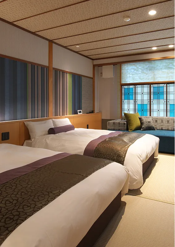 和の趣があるツインベッドルーム。畳の上に2台のベッドが置かれ、窓際には青いチェック柄の組子細工のような窓枠と、鮮やかなクッションが置かれたソファがある。