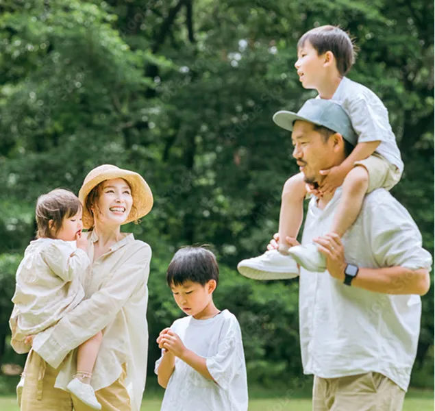 緑豊かな公園の芝生の上に立つ、4人家族のポートレート。父親が男の子を肩車し、母親が女の子を抱っこしている。