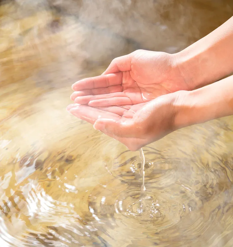 湯気が立ち上る温泉の湯を手で掬い上げているクローズアップ。指の間から湯が流れ落ち、湯面に波紋が広がっている。