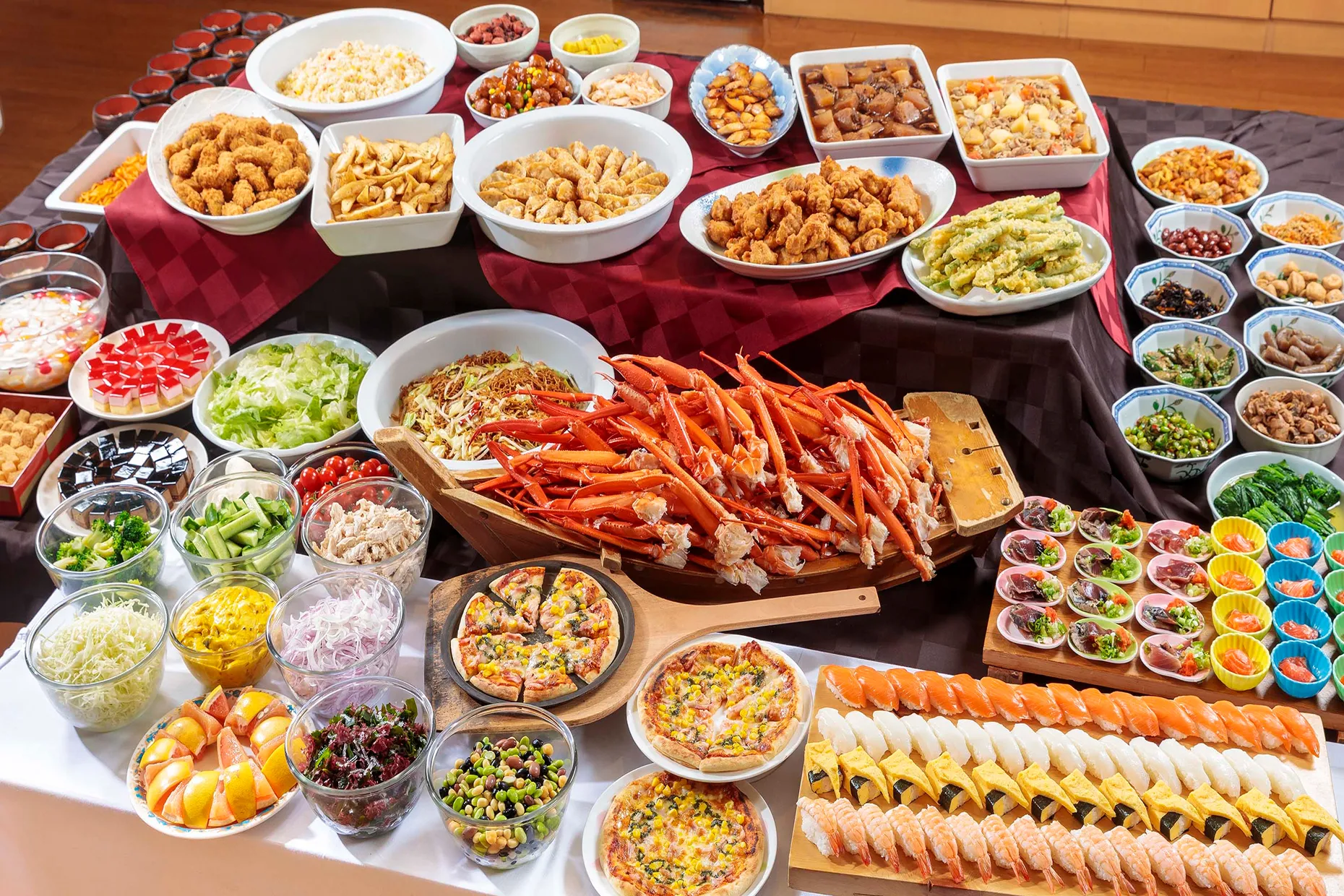 赤いクロスが敷かれた豪華なビュッフェ台に、山盛りのカニの脚、色鮮やかな握り寿司、ピザ、揚げ物、サラダ、フルーツ、そして小鉢に分けられたデザートまで、和洋中の料理が所狭しと並んでいる様子。