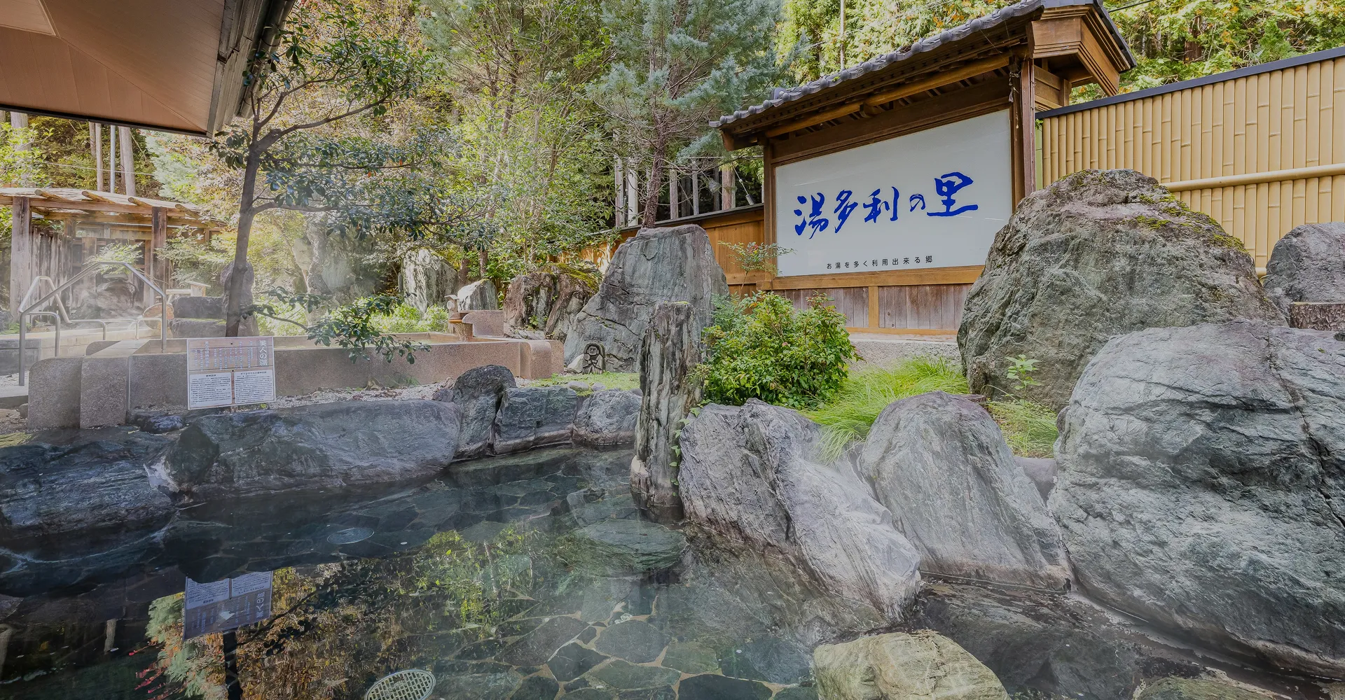 自然に囲まれた露天風呂の風景。手前にはゴツゴツとした岩に囲まれた、透明感のある湯船があり、奥には「湯めぐりの里」と書かれた木製の看板が見えます。周囲は豊かな緑と木々に囲まれ、自然の景観を活かした静かで落ち着いた造りになっており、ゆったりと温泉を楽しめる旅館の「湯」の素晴らしさを伝えています。