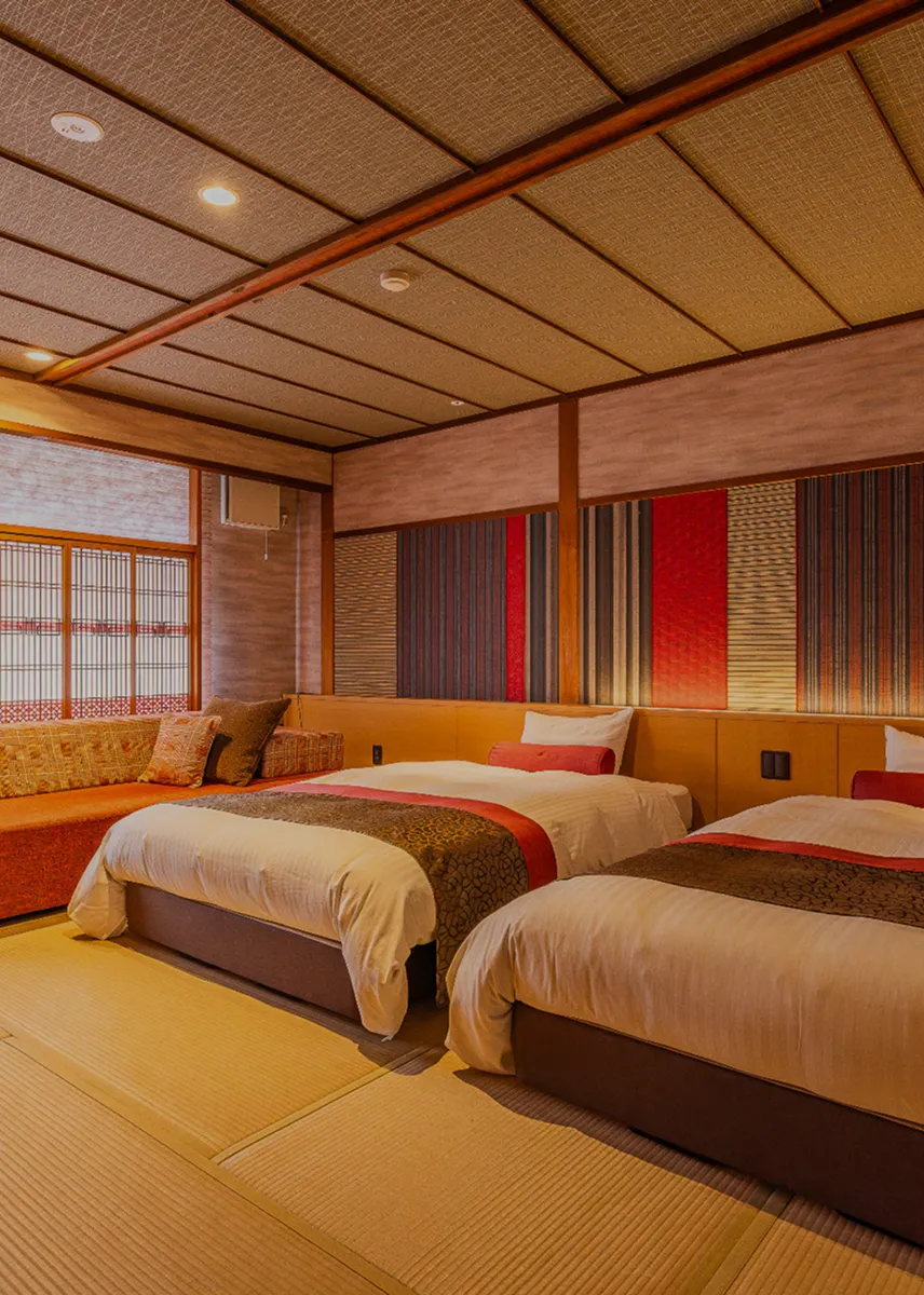 モダンなツインベッドルーム。部屋の床は畳敷きで、和の趣と洋の快適さを融合させた内装が特徴です。奥には和紙のようなブラインドがかかった窓と、オレンジ色のソファが配置され、くつろげる空間が提供されています。ベッドボードの背後には、赤や茶色を基調とした縞模様のアクセントウォールが施されており、落ち着きと華やかさを兼ね備えた旅館の「客室」の魅力が伝わります。