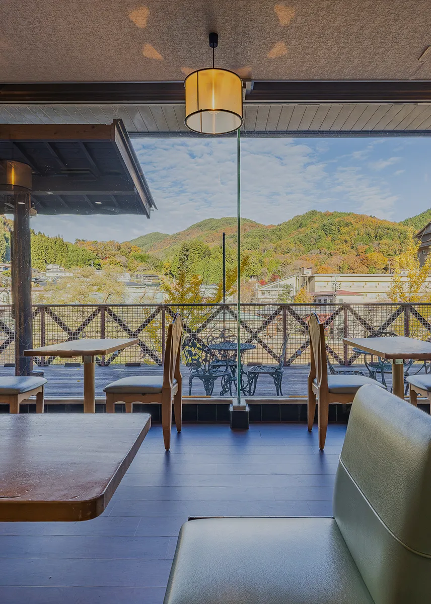 明るく開放的なラウンジ。大きな窓ガラスの向こうには、山々の緑と紅葉、そして温泉街の建物が見渡せます。室内には、木目のテーブルや椅子、ソファが配置され、吊り下げられた円形の照明が温かい雰囲気を作り出しています。美しい景観を眺めながら食事や休憩を楽しめる、旅館の「体験」空間の魅力が伝わります。