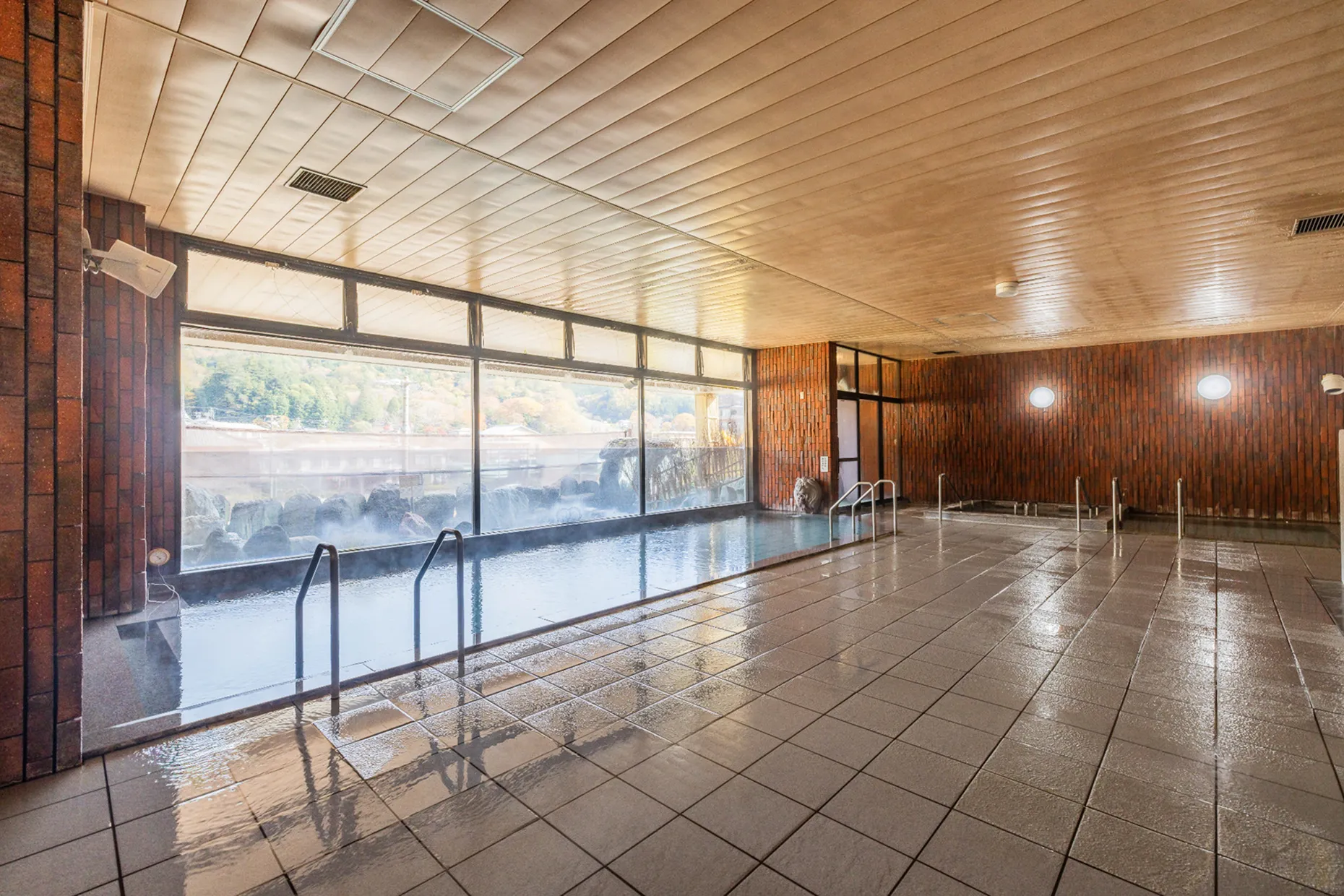 天井が高く広々とした温泉の大浴場。床と浴槽の縁は淡いベージュのタイル張りで、大きな窓沿いに細長い内湯がある。窓の外には露天風呂とその先に温泉街の景色が見える。
