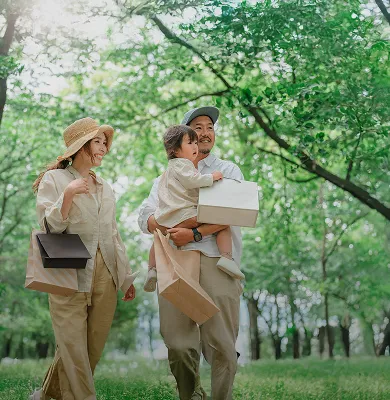 木々の緑豊かな公園を歩く、ベージュ系の服を着た3人家族。父親が幼い子どもを抱き、皆が紙袋を持っている。