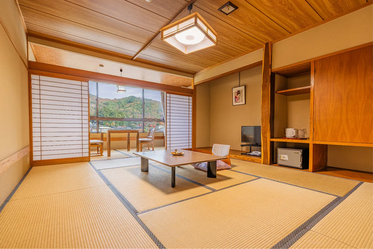 明るい日差しが差し込む14畳の純和室。手前には大きな座卓、奥の広縁（ひろえん）には椅子とテーブルが置かれ、大きな窓からは紅葉に彩られた山々の景色を一望できます。