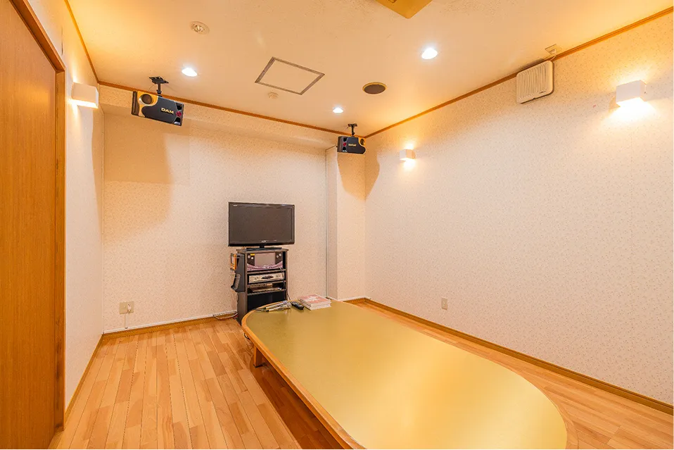 明るい木目調のフローリングが特徴的なカラオケルーム。中央には長い楕円形のテーブルがあり、モニターやカラオケ機材、天井設置のスピーカーが完備された、少人数からグループまで楽しく過ごせるプライベート空間です。