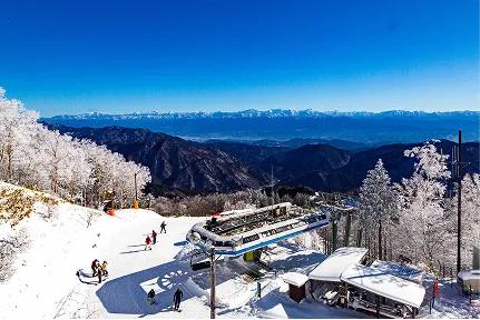 雪に覆われた山頂からロープウェイが伸び、青空の下でスキー客が冬の景色を楽しんでいる風景